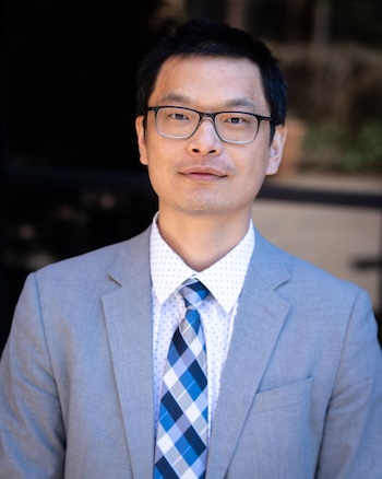 Mo Chen smiling at the camera with a grey suit and blue plaid tie.